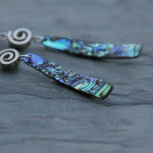 Può includere: Orecchini pendenti con rettangoli di conchiglia di abalone iridescente nei toni del blu, verde e viola. Gli orecchini hanno dettagli a spirale color argento. Sfondo grigio scuro.
