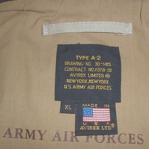 May include: A black label with white text reads "TYPE A-2, DRAWING NO. 30-1415, CONTRACT NO.1978-01, AVIREX LIMITED, NEW YORK, NEW YORK, U.S. ARMY AIR FORCES". A smaller label with white text reads "MADE IN" and features a red, white, and blue American flag. The label also has the text "XL, US. A. &reg;, AVIREX LTD.". The labels are attached to a tan fabric background with the text "ARMY AIR FORCES" printed in brown.