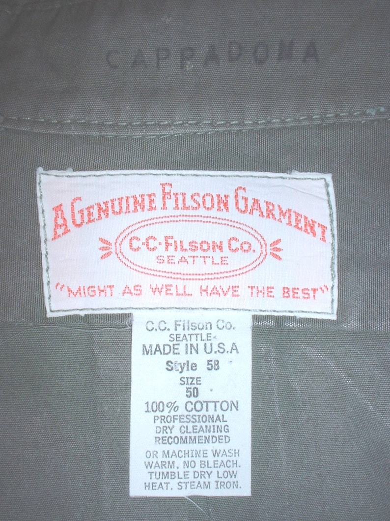 Op de afbeelding: Close-up van een kledinglabel met de tekst "A Genuine Filson Garment" en "C.C. Filson Co. Seattle". Het label bevat ook wasvoorschriften en de tekst "Made in U.S.A". Het kledingstuk is olijfgroen.