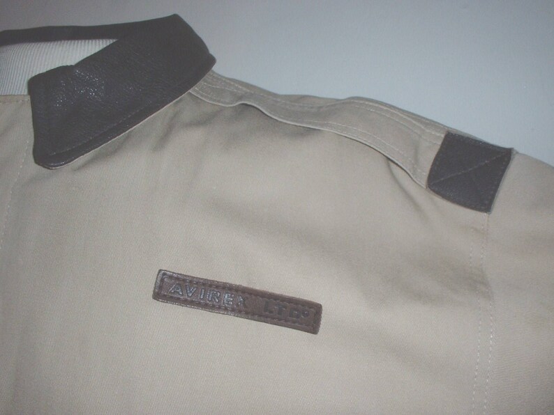 May include: A beige jacket with a brown leather collar and a brown leather patch on the shoulder. The patch has the word "AVIREX" on it.