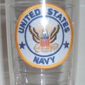 Puede incluir: Un vaso de plástico transparente con un parche de la Marina de los Estados Unidos en oro y azul. El parche presenta un águila calva con un escudo y un ancla.