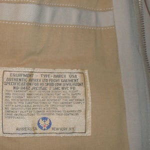 May include: A beige flight jacket label with the text "EQUIPMENT - TYPE - AVIREX USA" and "AUTHENTIC AVIREX LTD FLIGHT GARMENT" and "SPECIFICATION FOR HI SPEED LOW LEVEL FLIGHT" and "NO-8462 JRC35AC 2 SMC NYC PB". The label also features a small image of the Air Force emblem.