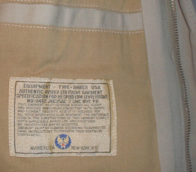 May include: A beige flight jacket label with the text "EQUIPMENT - TYPE - AVIREX USA" and "AUTHENTIC AVIREX LTD FLIGHT GARMENT" and "SPECIFICATION FOR HI SPEED LOW LEVEL FLIGHT" and "NO-8462 JRC35AC 2 SMC NYC PB". The label also features a small image of the Air Force emblem.