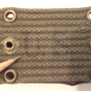May include: Close-up of a section of an olive green canvas belt. The belt has several evenly spaced metal grommets and a metal button. A pencil points towards one of the grommets.