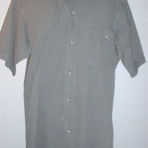 Op de afbeelding: Olijfgroen overhemd met korte mouwen, kraag, borstzak en knoopsluiting. Het overhemd heeft een klassiek ontwerp met epauletten op de schouders. De stof lijkt duurzaam, mogelijk een katoenmix, voor dagelijks gebruik.