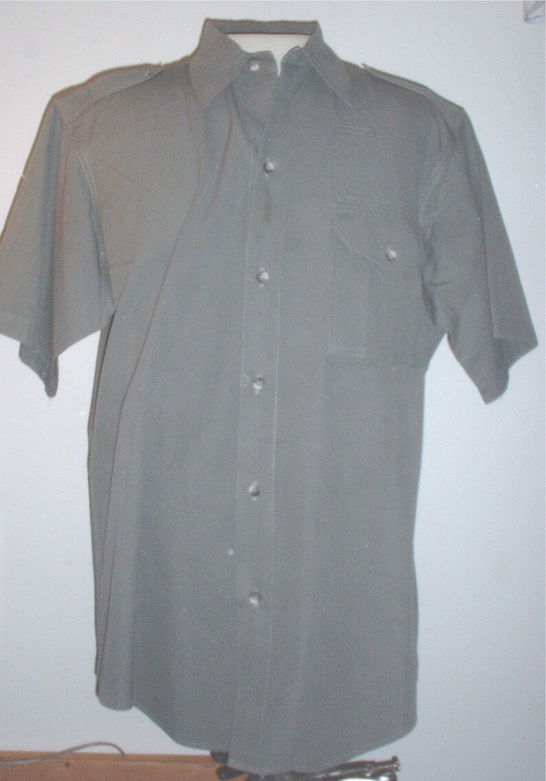 Op de afbeelding: Olijfgroen overhemd met korte mouwen, kraag, borstzak en knoopsluiting. Het overhemd heeft een klassiek ontwerp met epauletten op de schouders. De stof lijkt duurzaam, mogelijk een katoenmix, voor dagelijks gebruik.