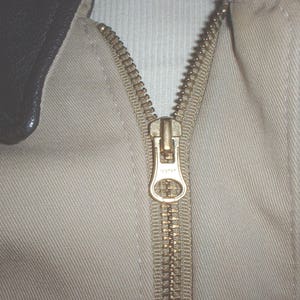 May include: A close-up of a beige jacket with a brown leather collar and a gold zipper. The zipper is pulled up and the jacket is closed.