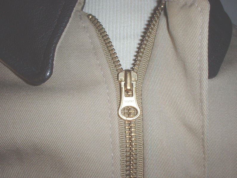 May include: A close-up of a beige jacket with a brown leather collar and a gold zipper. The zipper is pulled up and the jacket is closed.