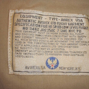 May include: A white tag with black text that reads "EQUIPMENT - TYPE - AVIREX USA AUTHENTIC AVIREX LTD FLIGHT GARMENT SPECIFICATION FOR HI SPEED LOW LEVEL FLIGHT NO:8462 JRC35AC 2 SMC NYC PB THIS GARMENT MUST BE WORN DURING ALL FLIGHT AND GROUND MANUOVERS CONSISTENT WITH SAFETY AND COMBAT SECURITY, ALSO IT IS REQUIRED FOR ALL HIGH SPEED VEHICULAR MOVEMENT THE MATERIALS USED IN THE CONSTRUCTIONS OF THIS GARMENT COMPLY WITH APPLICABLE AVIREX LTD SPECIFICATIONS NO SUBSTITUTES MAY BE ACCEPTED. GARMENT MUST BE CLEANED ACCORDING TO AVIREX LTD CARE INSTRUCTIONS TO PRESERVE THEIR CONTINUED USEFULNESS. AVIREXUSA NEW YORK NY". The tag also features a blue and white star emblem.