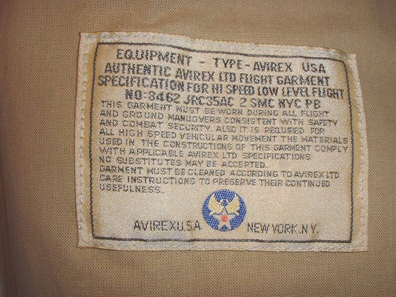 May include: A white tag with black text that reads "EQUIPMENT - TYPE - AVIREX USA AUTHENTIC AVIREX LTD FLIGHT GARMENT SPECIFICATION FOR HI SPEED LOW LEVEL FLIGHT NO:8462 JRC35AC 2 SMC NYC PB THIS GARMENT MUST BE WORN DURING ALL FLIGHT AND GROUND MANUOVERS CONSISTENT WITH SAFETY AND COMBAT SECURITY, ALSO IT IS REQUIRED FOR ALL HIGH SPEED VEHICULAR MOVEMENT THE MATERIALS USED IN THE CONSTRUCTIONS OF THIS GARMENT COMPLY WITH APPLICABLE AVIREX LTD SPECIFICATIONS NO SUBSTITUTES MAY BE ACCEPTED. GARMENT MUST BE CLEANED ACCORDING TO AVIREX LTD CARE INSTRUCTIONS TO PRESERVE THEIR CONTINUED USEFULNESS. AVIREXUSA NEW YORK NY". The tag also features a blue and white star emblem.