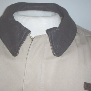 May include: A beige jacket with a brown leather collar. The jacket has a zip fastening and a patch on the lower right side.