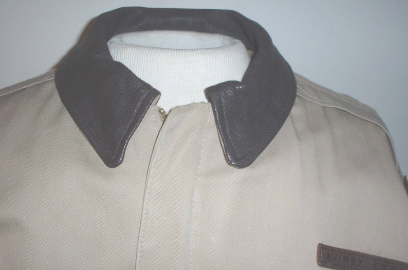 May include: A beige jacket with a brown leather collar. The jacket has a zip fastening and a patch on the lower right side.