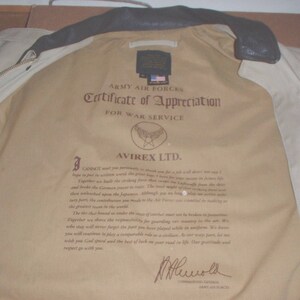 May include: A beige leather jacket with a certificate of appreciation from the Army Air Forces for war service. The certificate is signed by Avirex LTD. and includes a message of gratitude for service during World War II.