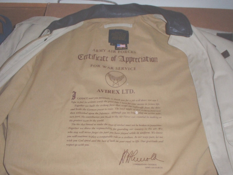 May include: A beige leather jacket with a certificate of appreciation from the Army Air Forces for war service. The certificate is signed by Avirex LTD. and includes a message of gratitude for service during World War II.