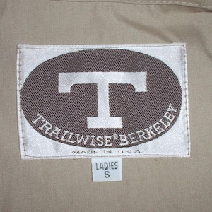 Könnte beinhalten: Ein weißes Etikett mit einem braunen Oval, das ein weißes großes T und den Text "TRAILWISE BERKELEY" in einem Kreis um das T enthält. Das Etikett trägt außerdem den Schriftzug "MADE IN U.S.A." und hat darunter ein kleineres weißes Etikett mit der Aufschrift "LADIES S".