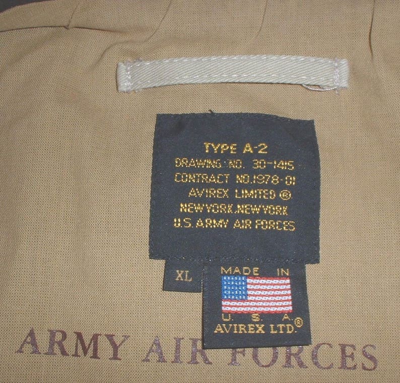 May include: A black label with white text reads "TYPE A-2, DRAWING NO. 30-1415, CONTRACT NO.1978-01, AVIREX LIMITED, NEW YORK, NEW YORK, U.S. ARMY AIR FORCES". A smaller label with white text reads "MADE IN" and features a red, white, and blue American flag. The label also has the text "XL, US. A. &reg;, AVIREX LTD.". The labels are attached to a tan fabric background with the text "ARMY AIR FORCES" printed in brown.