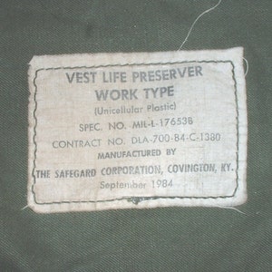 May include: A white label with black text that reads "VEST LIFE PRESERVER WORK TYPE (Unicellular Plastic) SPEC. NO. MIL-L-17653B CONTRACT NO. DLA-700-84-C-1380 MANUFACTURED BY THE SAFEGARD CORPORATION, COVINGTON, KY. September 1984".