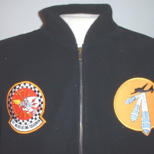 May include: Black fleece jacket with two embroidered patches. The left patch is a circular patch with a checkered background and the text "76 Air Refueling Squadron". The right patch is a circular patch with an orange background and a silhouette of a plane dropping bombs.