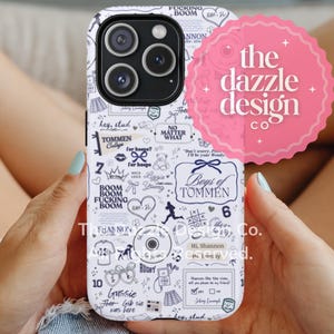 May include: A white iPhone case with a black and white doodle design, held by a person. The case features text and illustrations, including the words "BOOM," "TOMMEN," and "RUGBY." A pink circular logo with the text "the dazzle design co" is visible.