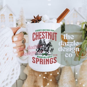 Puede incluir: Taza de cerámica blanca con el texto "SEASON'S GREETINGS FROM CHESTNUT SPRINGS" y un gráfico de una persona a caballo. La taza está llena de malvaviscos, una rama de canela y anís estrellado. La taza está sostenida por una persona.