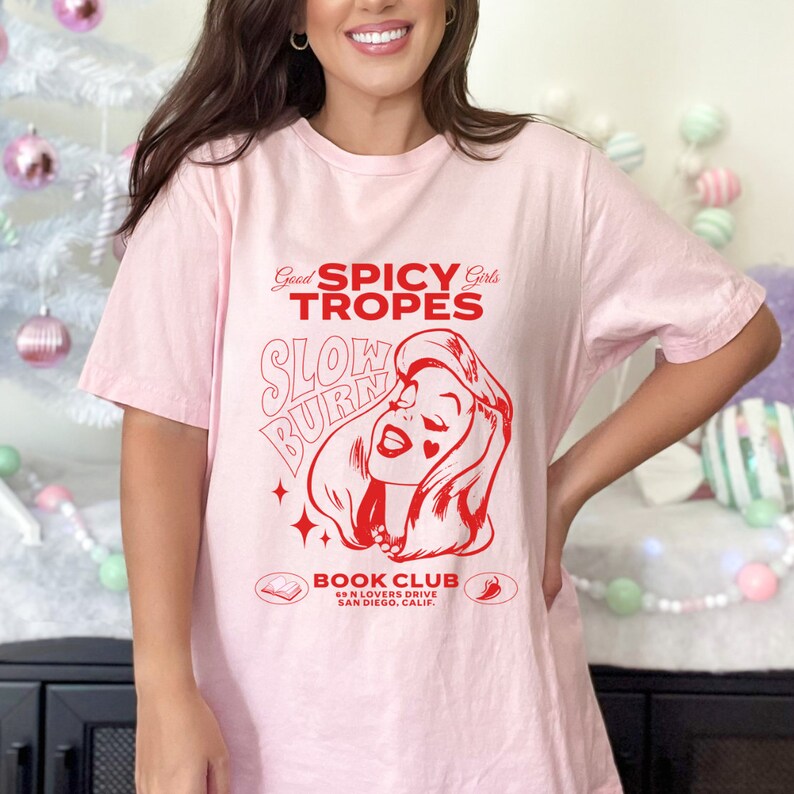 May include: Pink t-shirt with a red graphic design featuring a woman with the text "Good Spicy Girls Tropes Slow Burn Book Club 69 N Lovers Drive San Diego, Calif."