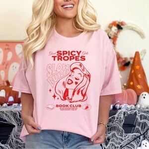May include: Pink t-shirt with a red graphic design that says "Good Spicy Girls Tropes Slow Burn" with a cartoon image of a woman. The text "Book Club 69 N Lovers Drive San Diego, Calif" is below the graphic.