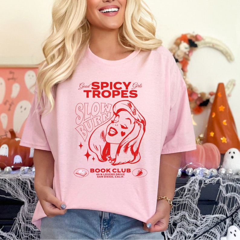 May include: Pink t-shirt with a red graphic design that says "Good Spicy Girls Tropes Slow Burn" with a cartoon image of a woman. The text "Book Club 69 N Lovers Drive San Diego, Calif" is below the graphic.