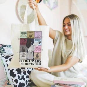 May include: A white tote bag with a colorful design featuring different book tropes. The bag has the text "Book Lovers The Tropes Tour" printed on the bottom.