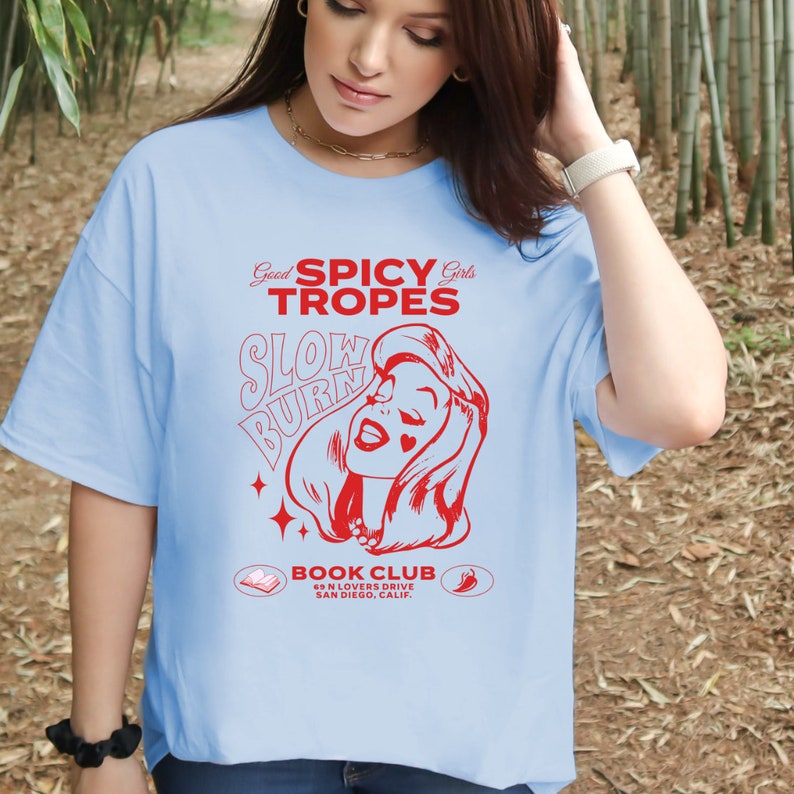 May include: A light blue t-shirt with a red graphic design featuring a woman with the text "Good Spicy Girls Tropes Slow Burn" and "Book Club 6N Lovers Drive San Diego, Calif."