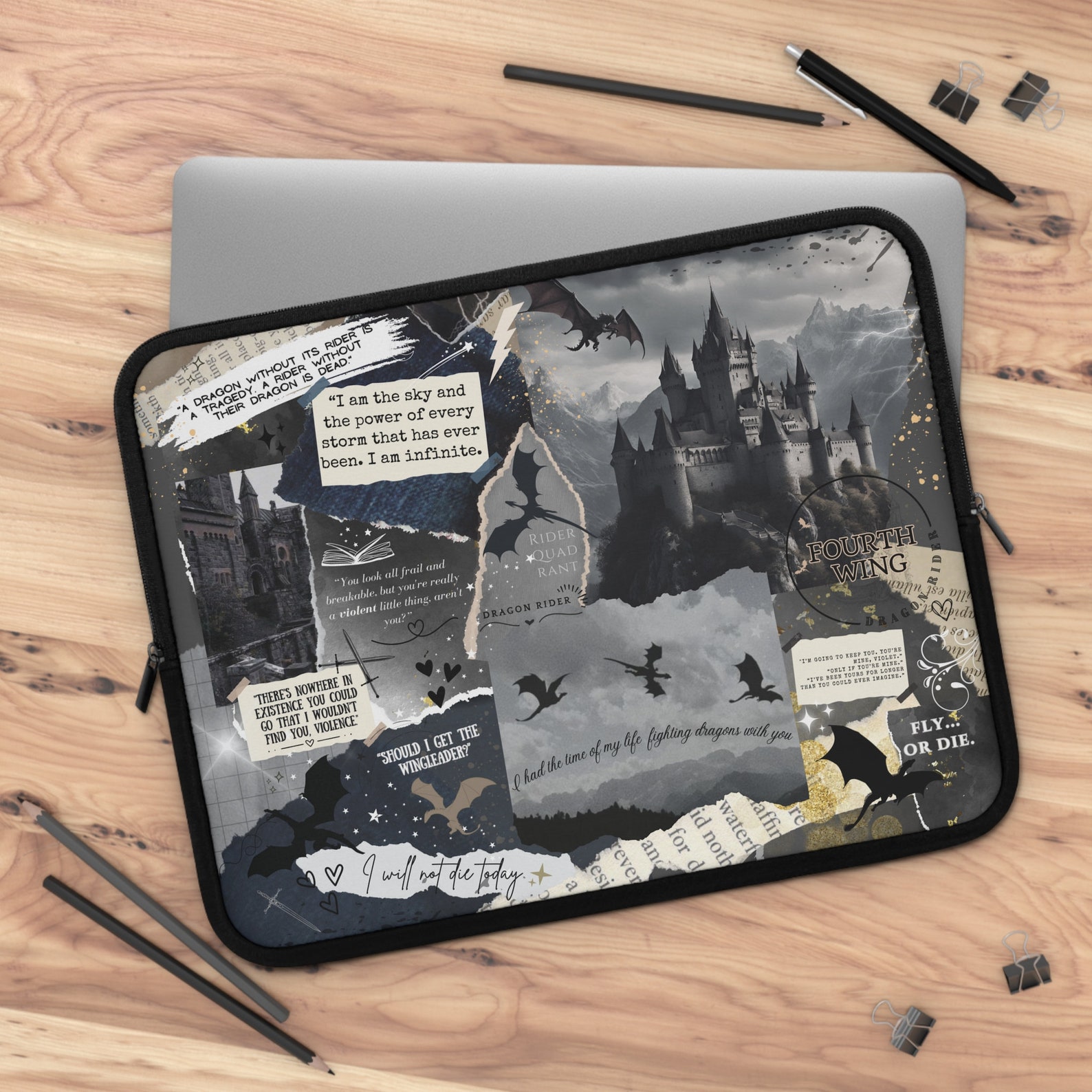 A light wooden background with a silver laptop on it. On top of that is a laptop case with a black, gray, and gold scrapbooking design with pictures of dragons, castles, and quotes from Fourth Wing.