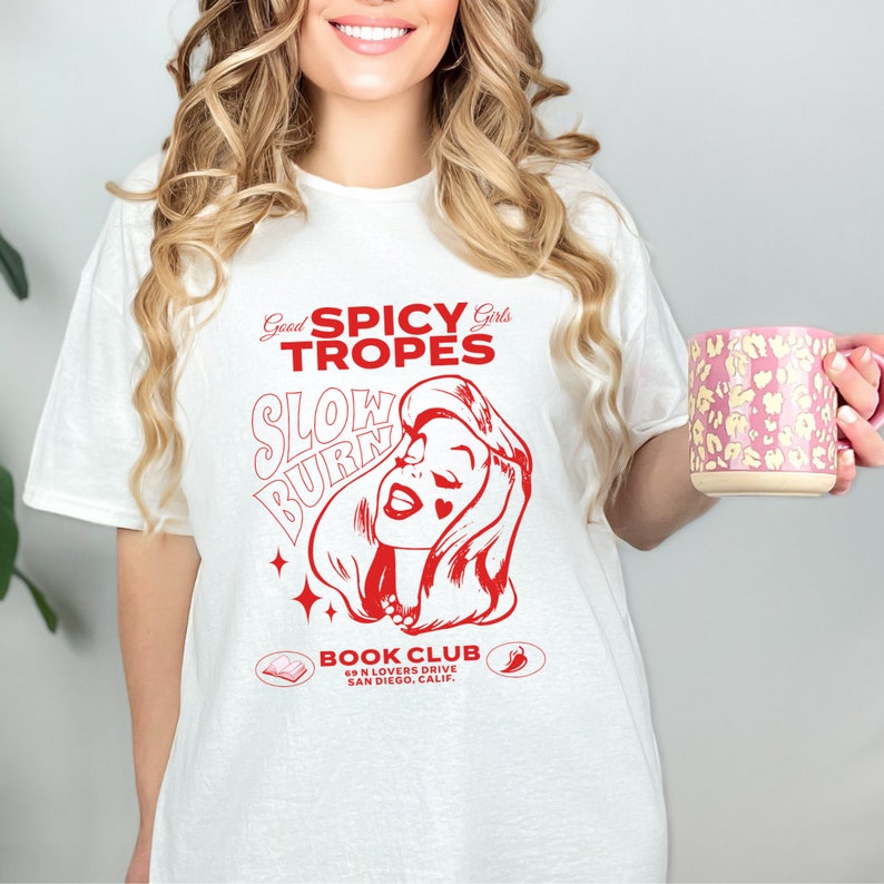May include: White t-shirt with a red graphic design that says "Good Spicy Girls Tropes Slow Burn" with a cartoon image of a woman with a red heart over her eye. The graphic also includes a book and a chili pepper with the text "Book Club 69 N Lovers Drive San Diego, Calif."