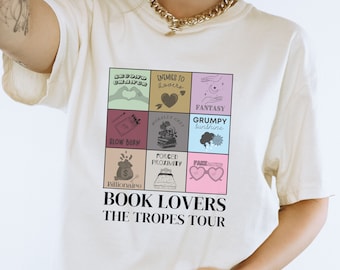 May include: A white t-shirt featuring a graphic print of a nine-square grid. Each square depicts a different book trope, including "Second Chance", "Enemies to Lovers", "Slow Burn", "Forced Proximity", "Billionaire", "Morally Grey", "Grumpy Sunshine", "Fantasy", and "Fake Dating". The text "BOOK LOVERS THE TROPES TOUR" is printed beneath the grid.