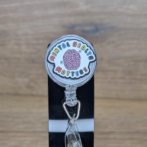 May include: A badge reel with a clear plastic clip and a round design. The design features the words "MENTAL HEALTH MATTERS" in rainbow colours around a pink brain illustration.