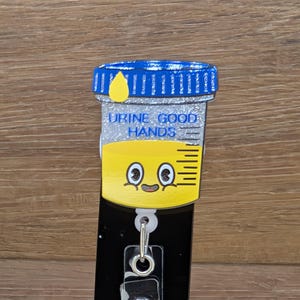 May include: A novelty badge reel with a cartoon urine sample container design. The container has a blue lid, yellow liquid, and the text "URINE GOOD HANDS." It features a smiling face and a clear plastic clip with a metal snap.