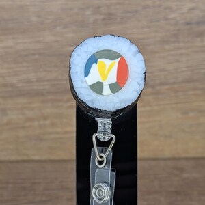 May include: A retractable badge reel with a sushi roll design. The reel has a black base and a clear plastic strap with a metal clip. The sushi roll has a white exterior and a colourful centre with blue, yellow, red, and green accents.