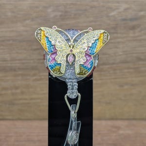May include: A butterfly-shaped badge holder. The wings are gold-coloured with colourful accents and rhinestones. The holder has a clear plastic strap and a metal clip. The badge reel is retractable.