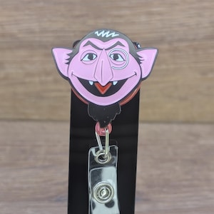 May include: A badge reel with a cartoon vampire character with a pink face, black hair, and a red collar. The reel has a clear plastic clip and a retractable cord. The character has a wide smile.