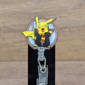 May include: A yellow Pikachu character wearing glasses, a black robe, and a red and gold scarf, attached to a retractable badge reel. The reel has a clear plastic strap and a metal clip. The badge reel is on a black background.