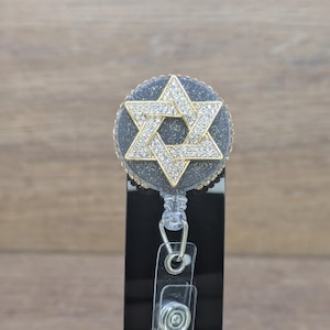 May include: A badge reel featuring a gold-coloured Star of David, embellished with rhinestones. The reel has a black background with gold glitter and a clear plastic clip.