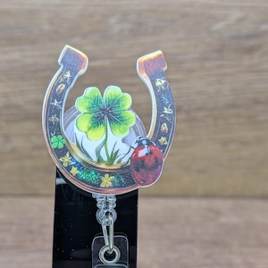 May include: A retractable badge reel with a horseshoe design, a four-leaf clover, and a ladybug. The horseshoe is brown with gold accents, and the clover is green. The reel has a clear plastic clip.