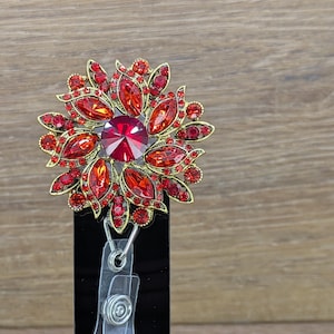 May include: A red and gold rhinestone flower badge reel. The flower design features a large, faceted red centre stone surrounded by smaller red rhinestones and gold-toned metal accents. The reel has a clear plastic strap and a black backing.