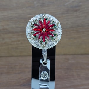 May include: A decorative badge reel featuring a poinsettia design. The center of the poinsettia is a deep red, surrounded by green leaves and clear rhinestones. The reel has a silver-colored, scalloped edge and a clear plastic clip.