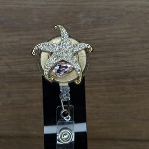 May include: A retractable badge holder featuring a gold-coloured starfish design adorned with clear rhinestones and a pink gemstone centre. The badge reel has a black plastic backing and a metal clip.