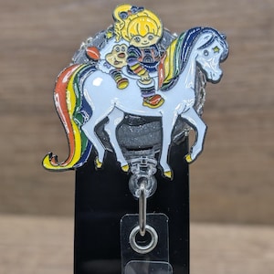 May include: A badge reel showing a cartoon character on a white unicorn with a rainbow mane and tail. The character wears colourful clothes. The reel has a black base and a retractable cord.