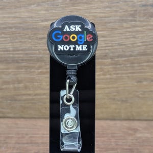 May include: A black retractable badge reel with the text "ASK Google NOT ME" in white, blue, red, and yellow. The reel has a clear plastic clip and a silver-coloured metal clasp.