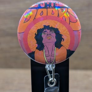 May include: A circular badge reel featuring a holographic finish and an illustration of a person with dark hair, wearing an orange top with a yellow neckline. The text "THE DOORS" is printed in large orange letters at the top of the circular design.