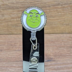 May include: A green Shrek face badge reel with a clear plastic retractable cord and a metal clip. The Shrek image is centered on a white background, surrounded by a clear, circular border. The reel is attached to a black, rectangular base.