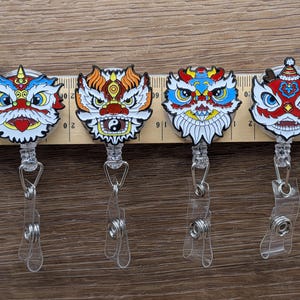 May include: Four colourful retractable badge holders with lion-like designs. Each badge reel has a different colour scheme, including red, blue, orange, and white, with intricate patterns. The badge reels have a clear plastic strap and a metal clip.
