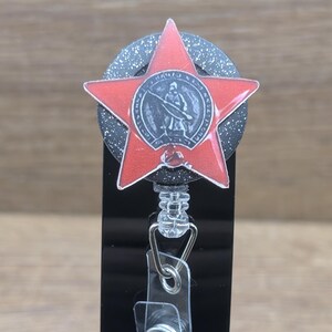 May include: A red star-shaped badge reel with a detailed emblem in the center. The badge reel has a black and silver glittery background and a clear plastic clip. The design is set against a blurred wooden background.