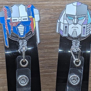 以下が含まれることがあります： エナメル製のキャラクターヘッドが付いたバッジリール2個。左側は青、赤、黄色のロボットの頭部、右側はグレー、白、紫のロボットの頭部がデザインされています。どちらも黒いプラスチック製の本体に、透明なビニールストラップと金属製のスナップボタンが付いています。